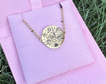 May include: A gold-colored necklace with a circular pendant featuring a castle design. The pendant is connected to a delicate chain and rests on a pink fabric. The necklace is displayed in a pink case, with green grass in the background.