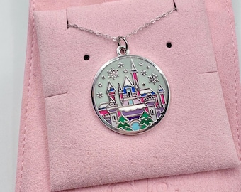 May include: A silver-toned necklace with a round pendant featuring a colorful castle design. The pendant has a light green background with snowflakes and a detailed illustration of a castle in shades of pink, blue, and purple. The necklace is displayed on a pink velvet background.