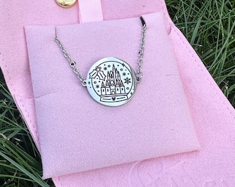 May include: A silver-colored necklace with a circular pendant featuring a castle design within a snow globe. The necklace is displayed on a pink pouch with a gold snap closure, set against a backdrop of green grass.