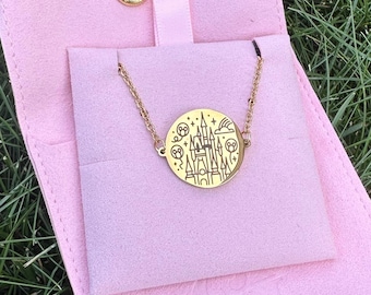May include: A gold-colored necklace with a circular pendant featuring a castle design, stars, and a rainbow. The necklace is displayed on a pink pouch with a gold snap closure, set against a backdrop of green grass.
