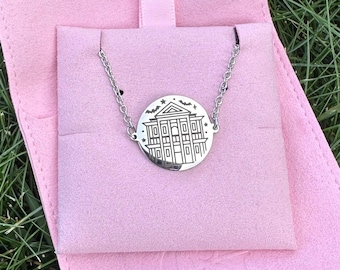 May include: A silver-colored necklace with a circular pendant featuring a detailed architectural design. The pendant is connected to a delicate chain. The necklace rests on a pink fabric, with a gold snap closure visible.