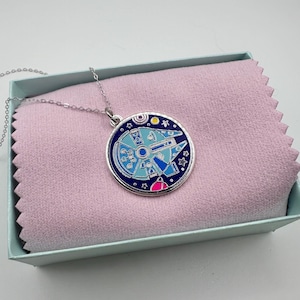 May include: A silver-toned necklace with a round pendant featuring a blue and silver spaceship design. The pendant has a dark blue border and is set against a pink background inside a light blue box. The necklace is a delicate chain.