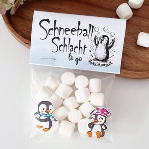 Könnte beinhalten: Ein klarer Beutel gefüllt mit weißen Marshmallows, mit einem Etikett mit der Aufschrift "Schneeball Schlacht to go" und einer Pinguin-Illustration. Zwei Pinguin-Aufkleber befinden sich auf dem Beutel. Der Beutel liegt auf einem Holztablett.