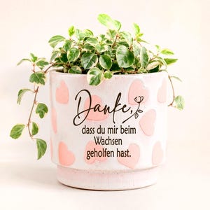 Könnte beinhalten: Ein weißer Keramik-Pflanztopf, verziert mit hellrosa Herzen und dem schwarzen Text "Danke, dass du mir beim Wachsen geholfen hast." Eine kleine grün-weiß panaschierte Pflanze mit hängenden Trieben wächst aus der Oberseite des zylindrischen Gefäßes.