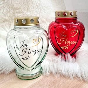 Könnte beinhalten: Zwei herzförmige Glas-Kerzenhalter. Einer ist klar, der andere ist rot. Beide haben goldfarbene Deckel und die Worte "Im Herzen nah" in Schrift. Der klare Kerzenhalter hat eine weiße Kerze.