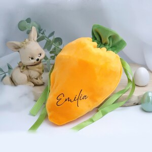 Könnte beinhalten: Eine orangefarbene Plüsch-Karotten-Kordeltasche mit grünem Oberteil und grünem Band. Die Tasche trägt den Namen "Emilia" in Schwarz.