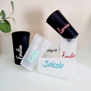 May include: A collection of personalized tumblers in black and frosted white. Each tumbler features a name in a different color: pink, teal, red, and white. The tumblers are arranged on a white surface.