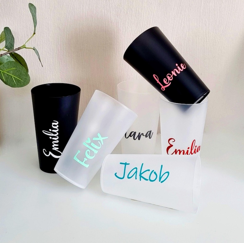 May include: A collection of personalized tumblers in black and frosted white. Each tumbler features a name in a different color: pink, teal, red, and white. The tumblers are arranged on a white surface.