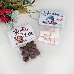 May include: Two clear plastic bags filled with treats. One bag contains brown chocolate-covered candies with a label reading Rentier Kacke and a reindeer illustration. The other bag contains white and pink marshmallows with a label reading Schneemann Pupse and a snowman illustration.