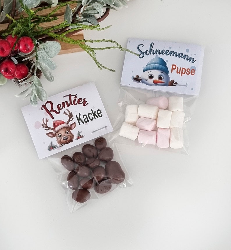 May include: Two clear plastic bags filled with treats. One bag contains brown chocolate-covered candies with a label reading Rentier Kacke and a reindeer illustration. The other bag contains white and pink marshmallows with a label reading Schneemann Pupse and a snowman illustration.