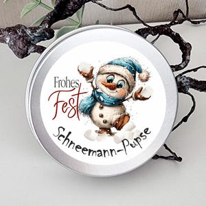 May include: A round silver metal tin featuring a circular label with a cartoon snowman wearing a blue hat and scarf, holding white marshmallows. The text reads Frohes Fest and Schneemann-Pupse in black and red script lettering on a white background.