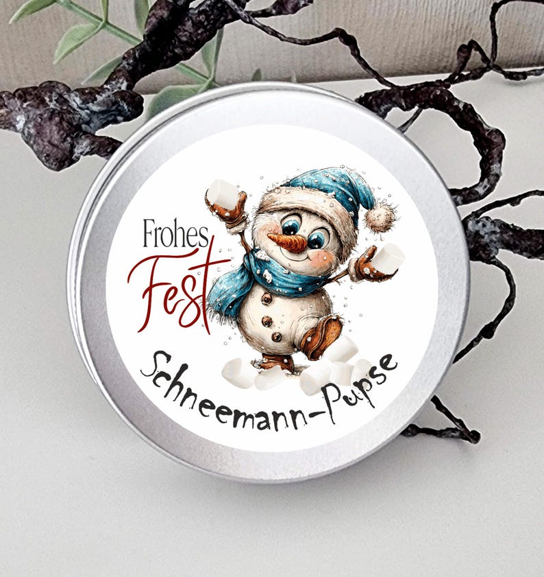 May include: A round silver metal tin featuring a circular label with a cartoon snowman wearing a blue hat and scarf, holding white marshmallows. The text reads Frohes Fest and Schneemann-Pupse in black and red script lettering on a white background.
