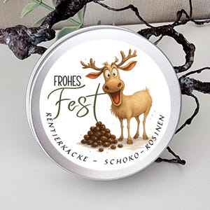 May include: A round silver metal tin featuring a cartoon reindeer with large eyes and antlers, a pile of brown chocolate-covered raisins, and the text "FROHES FEST RENTIERKACKE - SCHOKO - ROSINEN" printed in black and green lettering.
