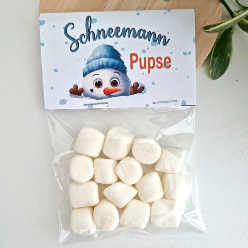May include: A clear plastic bag filled with white marshmallows, topped with a white paper header featuring a blue-hatted snowman illustration and the text "Schneemann Pupse" in blue and orange script and block lettering against a snowy background.