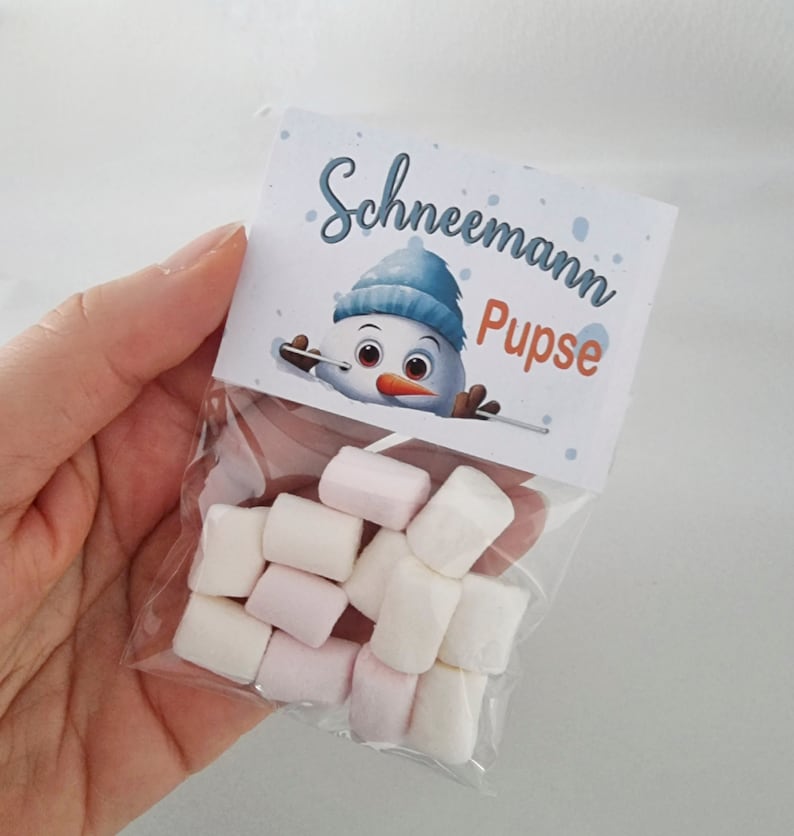 May include: A clear plastic bag filled with small, cylindrical white and light pink marshmallows. The bag is sealed with a white paper topper featuring a cartoon snowman wearing a blue hat and the text "Schneemann Pupse" in blue and orange.