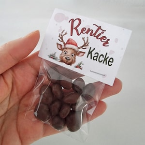 May include: A small clear plastic bag filled with dark brown chocolate-covered candies, topped with a white paper header card featuring a cartoon reindeer wearing a Santa hat and the printed text Rentier Kacke in dark red and black lettering.