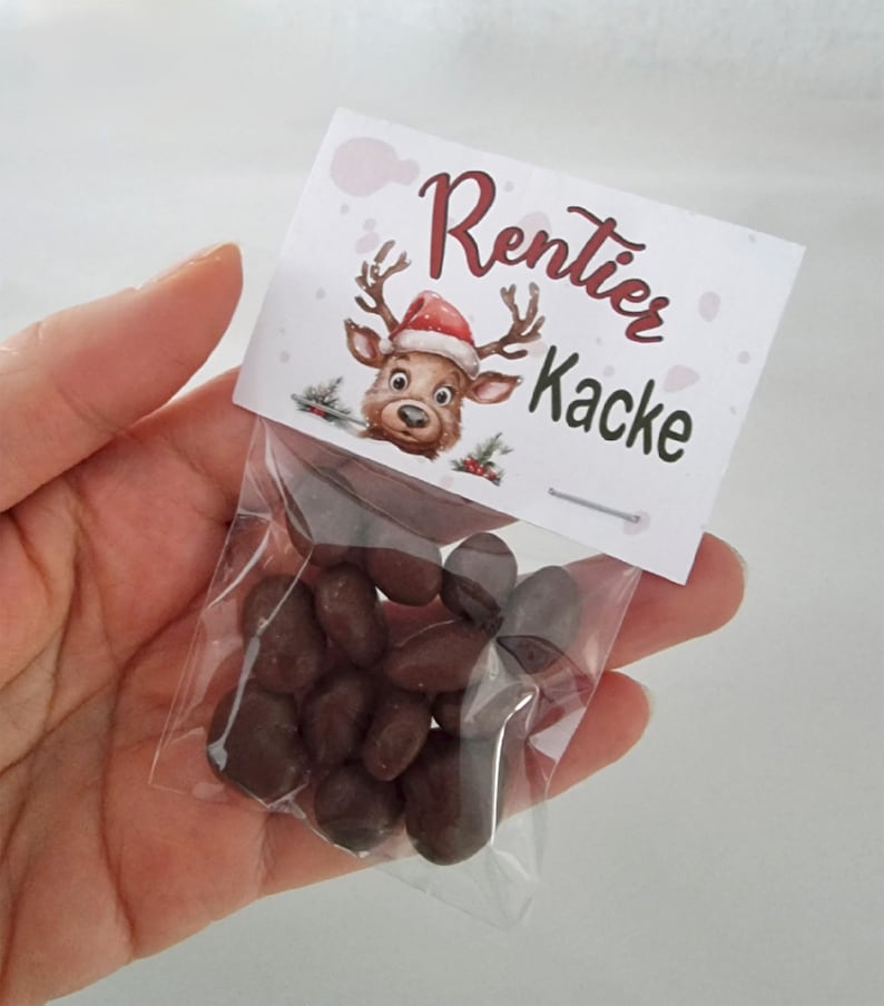 May include: A small clear plastic bag filled with dark brown chocolate-covered candies, topped with a white paper header card featuring a cartoon reindeer wearing a Santa hat and the printed text Rentier Kacke in dark red and black lettering.