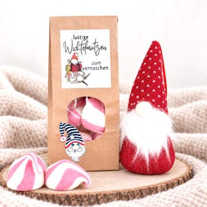 May include: A brown paper bag with a circular window containing pink and white striped marshmallows, labeled with the text "lustige Wichtelmützen zum vernaschen" and a gnome illustration, next to a red knit gnome with a white beard and two marshmallows.