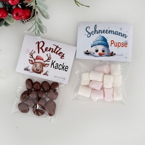 May include: Two clear plastic bags filled with treats. One bag contains brown chocolate-covered candies with a label reading Rentier Kacke and a reindeer illustration. The other bag contains white and pink marshmallows with a label reading Schneemann Pupse and a snowman illustration.