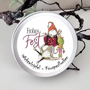 May include: A round silver metal tin featuring a label with the text Frohes Fest and Wichtelzipfel - Knusperflocken. The illustration shows two gnomes, one sitting in a large wine glass with string lights and another gnome lying below.