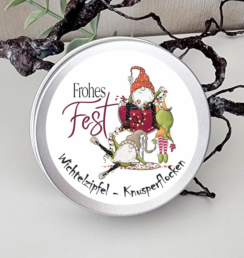 May include: A round silver metal tin featuring a label with the text Frohes Fest and Wichtelzipfel - Knusperflocken. The illustration shows two gnomes, one sitting in a large wine glass with string lights and another gnome lying below.