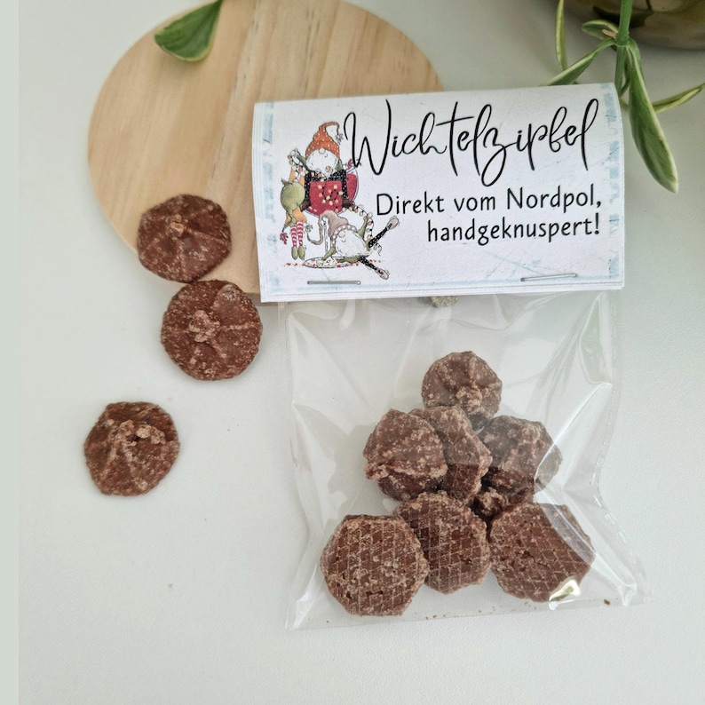 May include: A clear plastic bag containing small, round, brown chocolate cookies. The bag has a white label with a cartoon gnome illustration and the text "Wichtelzipfel Direkt vom Nordpol, handgeknuspert!". Three loose cookies sit outside the bag.