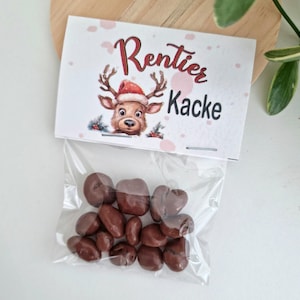 May include: A clear plastic bag filled with small, round, brown chocolate candies. The bag is sealed with a white paper header card featuring a cartoon reindeer wearing a red Santa hat and the printed text Rentier Kacke in red and black.
