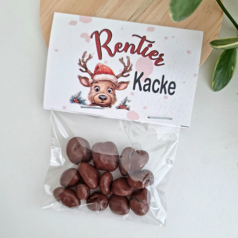 May include: A clear plastic bag filled with small, round, brown chocolate candies. The bag is sealed with a white paper header card featuring a cartoon reindeer wearing a red Santa hat and the printed text Rentier Kacke in red and black.