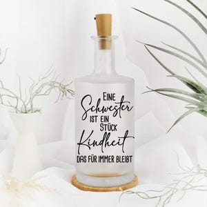 Könnte beinhalten: Eine frosted Glasflasche mit einem Korkverschluss und einem schwarzen Vinyl-Aufkleber mit der Aufschrift "Eine Schwester ist ein Stück Kindheit das für immer bleibt".