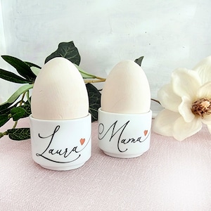 Könnte beinhalten: Zwei weiße Keramik-Eierbecher, einer mit dem Namen "Laura" und einem kleinen Herz, der andere mit dem Wort "Mama". Jeder Becher enthält ein helles Ei. Der Hintergrund umfasst Grün und eine weiße Blume.