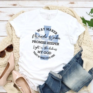 May include: White t-shirt with a blue cross design and the text "WAY MAKER Miracle Worker PROMISE KEEPER Light in the darkness MY GOD THAT IS WHO YOU ARE"