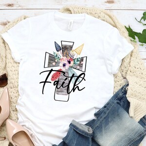 May include: White t-shirt with a floral design on a wooden cross. The word "Faith" is written in black script below the cross.
