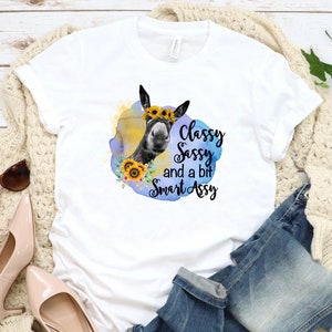 May include: White t-shirt featuring a graphic of a donkey wearing a sunflower crown, surrounded by a watercolor design. The text reads "Classy, Sassy, and a bit Smart Assy."