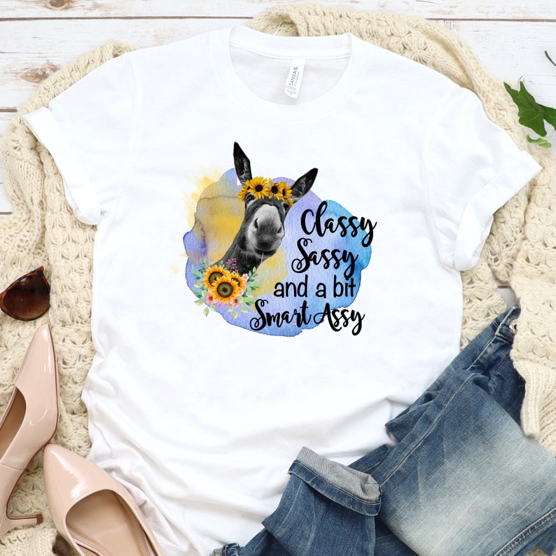 May include: White t-shirt featuring a graphic of a donkey wearing a sunflower crown, surrounded by a watercolor design. The text reads "Classy, Sassy, and a bit Smart Assy."