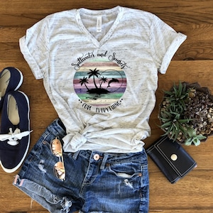 May include: A white t-shirt with a distressed look featuring a graphic of a sunset with palm trees and a dolphin. The text "Saltwater and Sunsets" is above the graphic and "Curl Everything" is below the graphic.