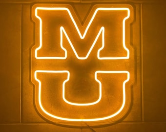 Könnte beinhalten: Ein Neon-Schild mit den Buchstaben "MU" in leuchtend gelber Farbe. Das Schild ist an einer Wand montiert.