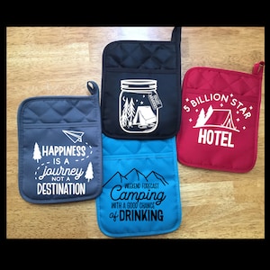 May include: Four different pot holders with a camping theme. The first pot holder is gray with the text "Happiness is a journey not a destination". The second pot holder is black with a mason jar design and the text "Collect Memories". The third pot holder is red with the text "5 Billion Star Hotel". The fourth pot holder is blue with the text "Weekend Forecast Camping with a good chance of drinking".