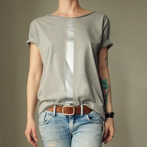 Peut inclure: Un t-shirt gris avec une bande argentée sur le devant. Le t-shirt est porté avec une ceinture en cuir marron et un jean bleu.