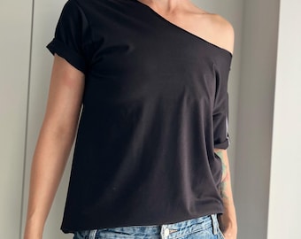 May include: Black off-the-shoulder t-shirt with short sleeves. The top has a unique asymmetrical neckline and a relaxed fit. The sleeves are rolled up. The model is wearing blue jeans.