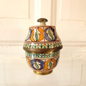 Könnte beinhalten: Eine dekorative Keramikdose mit Deckel, mit einem farbenfrohen Muster aus blauen, orangefarbenen und grünen Designs. Die Dose hat Messingakzente und einen Messingknopf oben. Die Dose ist etwa 13 cm hoch.