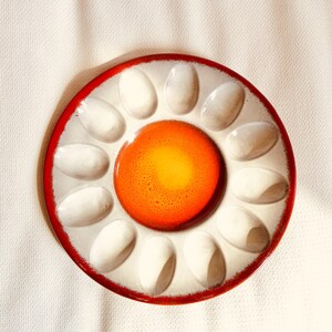 Könnte beinhalten: Ein runder Eierteller aus Keramik mit einer leuchtend orange-gelben Mitte, umgeben von zwölf weißen, ovalen Vertiefungen. Der Teller hat einen markanten roten Rand, der das gesamte Design einrahmt und eine blumenartige Optik erzeugt.