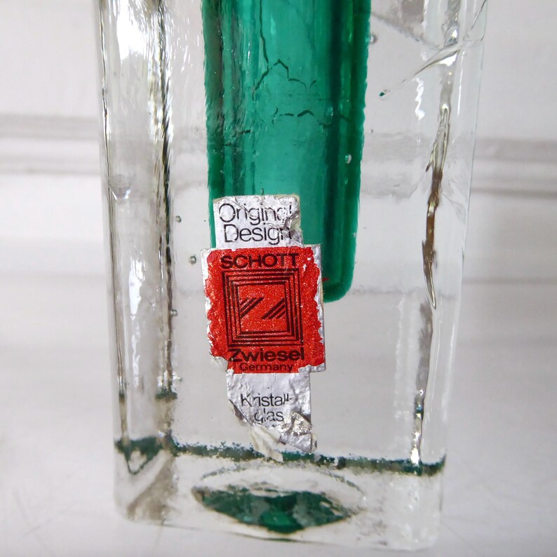 May include: Clear glass vase with a textured surface and a vibrant green interior. A red and white label with the text "Original Design SCHOTT Zwiesel Germany Kristall Glas" is affixed to the front. The vase has a rectangular shape.