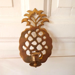 Puede incluir: Un aplique de pared en forma de piña de latón con un portavelas. El aplique tiene un diseño detallado con secciones recortadas y una superficie texturizada. El portavelas tiene forma de copa pequeña. El aplique está montado en una puerta blanca.