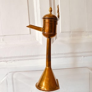 Puede incluir: Una lámpara de aceite antigua de latón, con un tallo alto y delgado y una base cónica. La lámpara presenta un pequeño pico, una tapa con bisagras y un remate decorativo. El latón tiene una pátina cálida y envejecida.