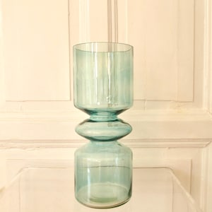Könnte beinhalten: Eine hohe, zylindrische Glasvase in einem durchscheinenden hellblauen Farbton. Die Vase zeichnet sich durch ein markantes, gestapeltes Design mit einem breiten Boden, einem kleineren, abgerundeten Mittelteil und einem geraden, oben offenen Zylinder aus.