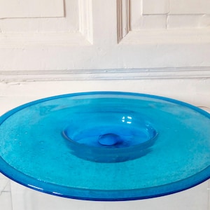Peut inclure: Un bol en verre turquoise vif avec un design circulaire. Le bol présente une zone centrale surélevée et un large bord plat. Le verre semble avoir une surface texturée, ajoutant un intérêt visuel à la pièce.