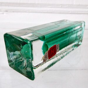 May include: A rectangular, clear glass bottle with a vibrant green hue. The bottle has a slightly textured surface and a red and white label. The bottle is lying on its side, showcasing its unique shape and color.