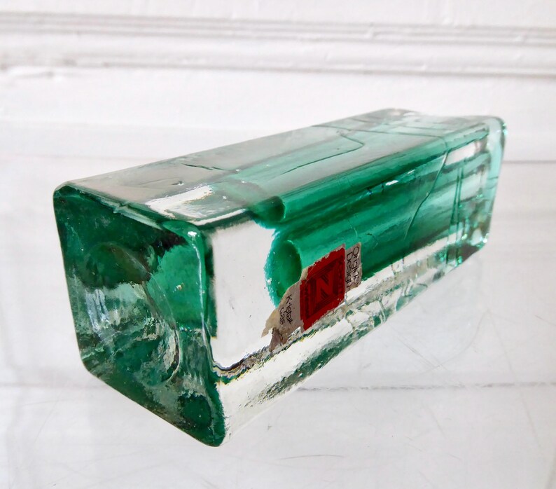 May include: A rectangular, clear glass bottle with a vibrant green hue. The bottle has a slightly textured surface and a red and white label. The bottle is lying on its side, showcasing its unique shape and color.