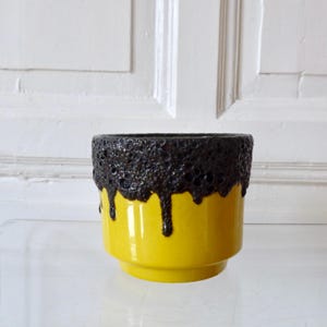 Puede incluir: Una maceta de cerámica con base amarilla brillante y borde marrón oscuro texturizado. El borde marrón tiene un efecto de goteo, agregando un elemento de diseño único. El objeto es decorativo, adecuado para interiores.