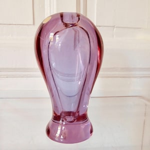 May include: A tall, elegant, lavender-colored glass vase with a rounded body and a flared base. The vase has a smooth, glossy finish and a unique, elongated shape. The vase is a decorative item.